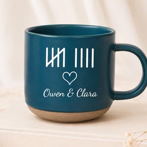 May include: A dark teal ceramic mug with a matte finish and a natural clay base. The front features white printed tally marks, a small heart icon, and the names Owen & Clara in a cursive script. The mug has a rounded handle on the right side.