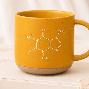 May include: A mustard yellow ceramic mug with a speckled finish and a white chemical structure of caffeine printed on the front. The mug features a rounded handle and a smooth, unglazed base, showcasing a clean and functional design for daily use.
