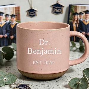 Könnte beinhalten: Eine rosa Keramiktasse mit dem Text "Dr. Benjamin EST. 2026" steht im Mittelpunkt. Die Tasse ist von Accessoires zum Thema Abschluss umgeben, darunter ein Fotobuch, Schlüsselanhänger und eine Halskette, alle mit einem Abschlusskappen-Design.