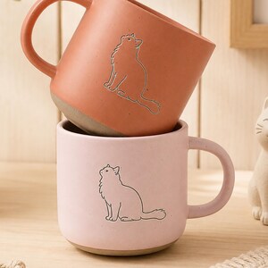 May include: Two ceramic mugs stacked vertically. The top mug is terracotta orange with a line drawing of a sitting cat. The bottom mug is light pink with a matching line drawing of a sitting cat. Both mugs feature a smooth finish and a handle.