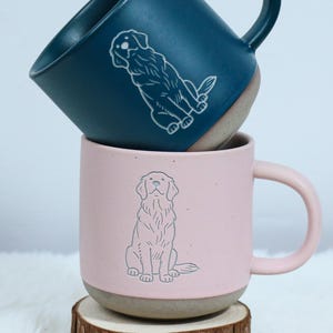 May include: Two ceramic mugs, one teal and one pink, stacked on a wooden coaster. Each mug features a white line drawing of a dog. The teal mug is on top, and the pink mug is below. The mugs have a neutral-coloured base and matching handles.