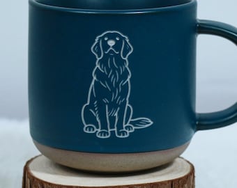 May include: A teal ceramic mug with a white line drawing of a dog. The mug has a brown base and a large handle. The mug is sitting on a small, round wooden slice.