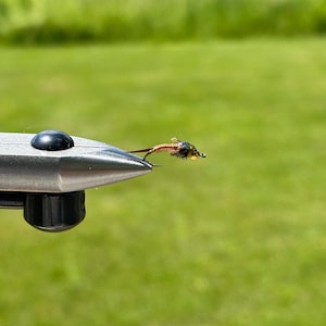 Könnte beinhalten: Eine kleine künstliche Fliege zum Angeln, die in einem Metallschraubstock eingespannt ist. Die Fliege hat einen mit Kupferdraht umwickelten Körper, einen dunklen Thorax mit feinen Fasern und einen goldfarbenen Perlenkopf.