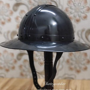 Puede incluir: Un casco de metal negro con un ala ancha y detalles remachados. El casco tiene una corona redondeada y una correa para la barbilla. La imagen incluye la palabra "Fotógrafo" y la fecha y hora de la foto.