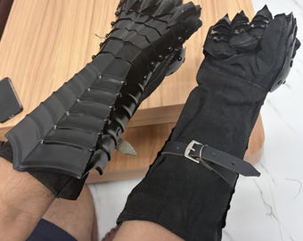 Könnte beinhalten: Ein Paar schwarze Handschuhe mit beweglichen Metallplatten auf dem Handrücken und den Fingern, befestigt an einem schwarzen Stoffärmel. Der Ärmel verfügt über einen verstellbaren schwarzen Lederriemen mit einer silberfarbenen Metallschnalle.