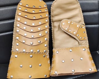 May include: A pair of tan leather mittens featuring layered, overlapping plates on the back of the hand and a separate thumb section. Both pieces are decorated with numerous silver-coloured metal studs and finished with visible whip-stitched edges.