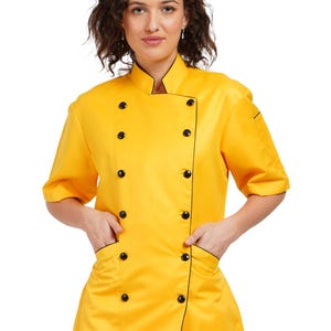 May include: A vibrant yellow chef's jacket with short sleeves and a mandarin collar. The jacket has black buttons and black trim along the edges, pockets, and cuffs. The model is wearing the jacket and has their hands in the pockets.