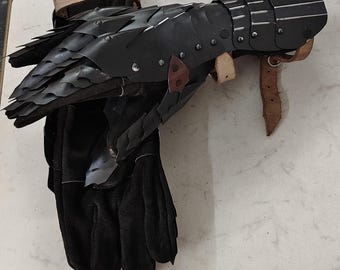 Könnte beinhalten: Ein schwarzer Metallhandschuh mit überlappenden, beweglichen Platten, die die Finger und den Handrücken bedecken, befestigt an einem schwarzen Wildlederhandschuh. Die Rüstung verfügt über silberfarbene Nieten und beige Lederriemen mit Metallschnallen.