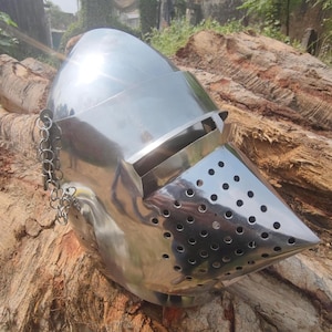 May include: A shiny, silver-coloured medieval helmet with a pointed visor and small ventilation holes. A chain is attached to the side. The helmet is resting on a weathered, brown wooden surface.