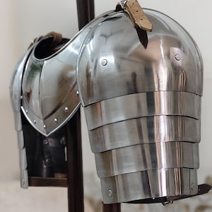 May include: A polished steel shoulder armor piece, showcasing a layered design for the arm and a curved plate for the shoulder. The armor is held in place by leather straps and rivets, reflecting light.