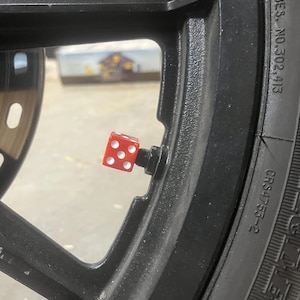 May include: A red dice valve stem cap on a black motorcycle wheel. The dice has white dots and is mounted on a black valve stem. The tire has the text "CRS4753-2".