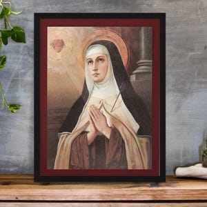 May include: Framed religious art print featuring a portrait of a nun with clasped hands. The artwork has a warm color palette with a black frame and a red inner border. The background includes a heart and a column.