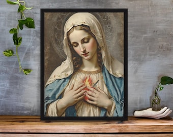 May include: Framed print of a religious artwork. The image depicts a woman with a serene expression, wearing a white veil and blue robe. Her hands are clasped over her chest, with a flaming heart visible. The artwork is framed in a black border.