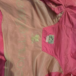 May include: Bright pink windbreaker jacket with a light pink and beige lining. The jacket has a zipper closure and a tag with the brand name. The interior lining features the word "conner" in a light green color.
