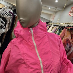 May include: A bright pink jacket with a hood and a white zipper. The jacket is displayed on a mannequin in a clothing store. The jacket has a sporty design and appears to be made of a lightweight material.