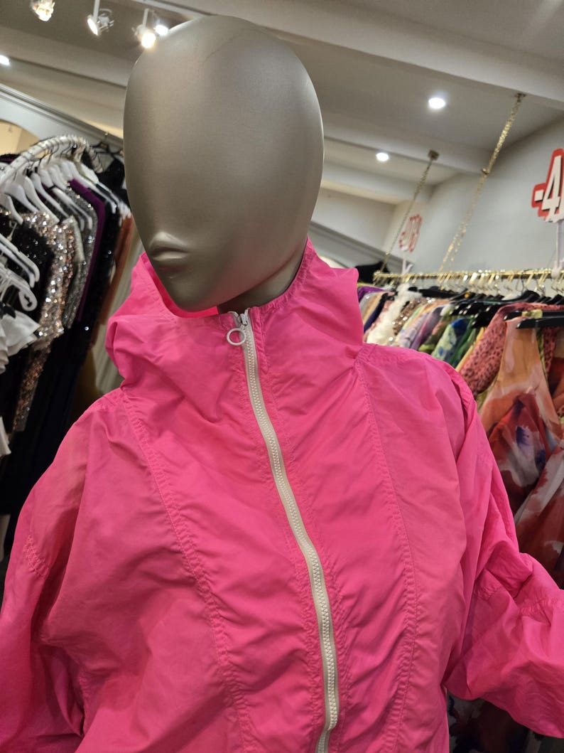 May include: A bright pink jacket with a hood and a white zipper. The jacket is displayed on a mannequin in a clothing store. The jacket has a sporty design and appears to be made of a lightweight material.