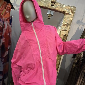 May include: Bright pink hooded jacket with a white zipper and drawstring. The jacket is displayed on a mannequin. The jacket has a casual style and is made of a lightweight material. The jacket is a vibrant color.