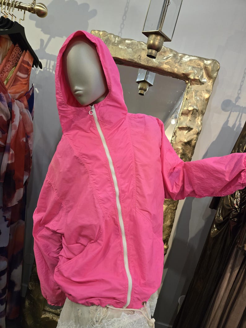 May include: Bright pink hooded jacket with a white zipper and drawstring. The jacket is displayed on a mannequin. The jacket has a casual style and is made of a lightweight material. The jacket is a vibrant color.