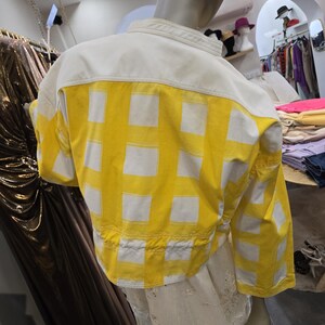 May include: A cropped jacket with a white upper back and sleeves, and a yellow lower back and sleeves. The yellow fabric features a white grid pattern. The jacket is displayed on a mannequin in a clothing store.