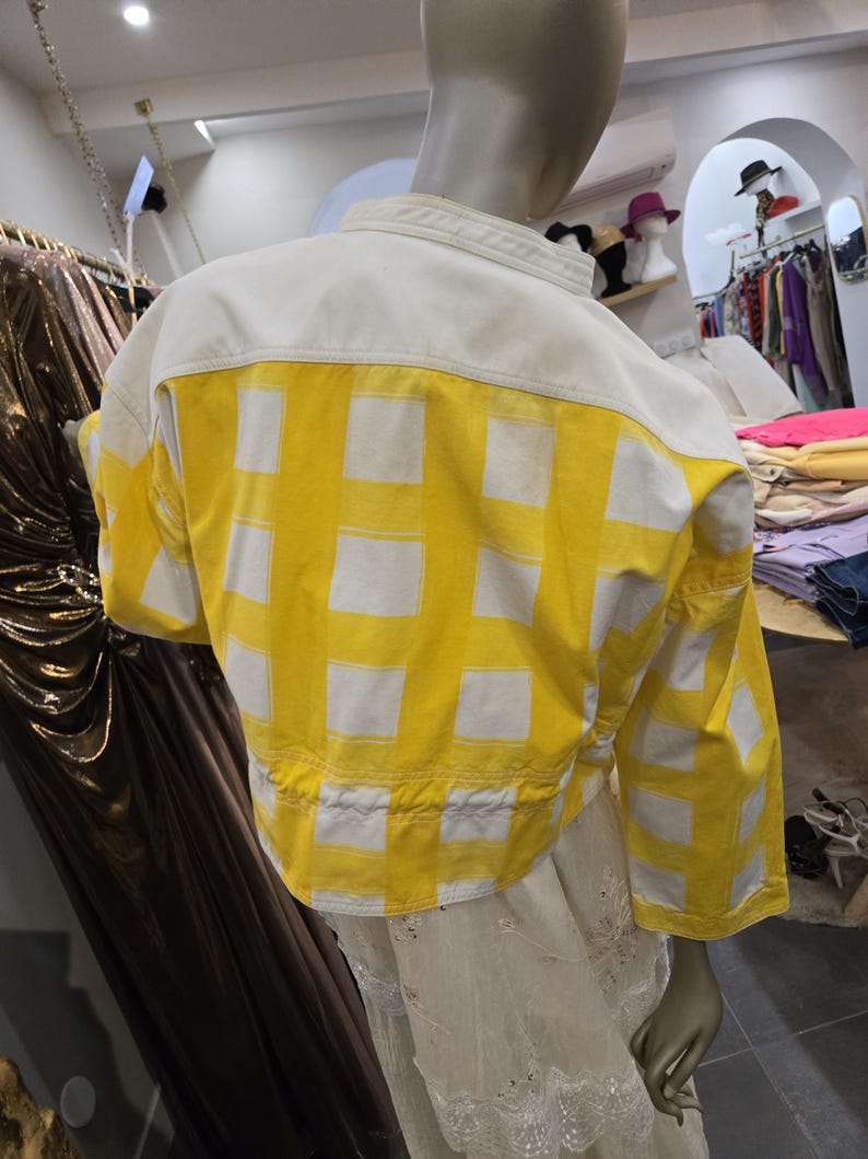 May include: A cropped jacket with a white upper back and sleeves, and a yellow lower back and sleeves. The yellow fabric features a white grid pattern. The jacket is displayed on a mannequin in a clothing store.