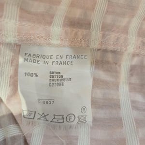 Może przedstawiać: Zbliżenie r&oacute;żowo-białej tkaniny w paski z metką. Na metce widnieje napis "FABRIQUE EN FRANCE MADE IN FRANCE" i wyszczeg&oacute;lniony skład tkaniny jako 100% bawełny w wielu językach. Widoczne są r&oacute;wnież instrukcje prania.