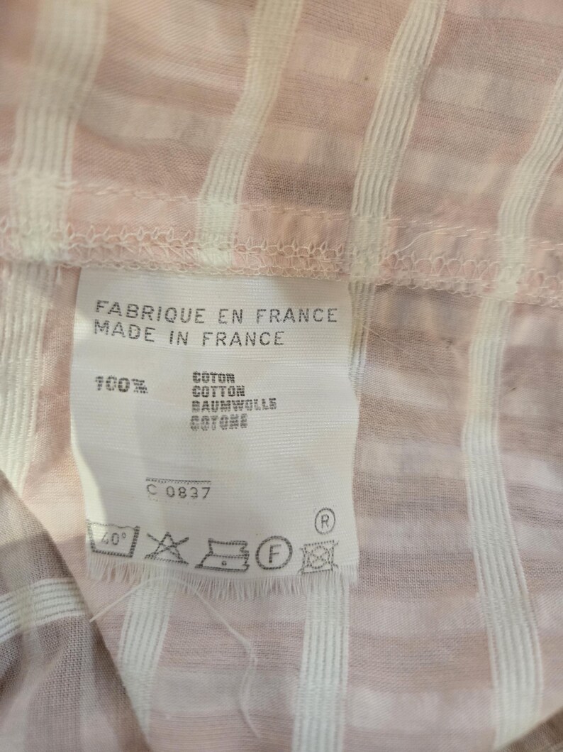 Może przedstawiać: Zbliżenie r&oacute;żowo-białej tkaniny w paski z metką. Na metce widnieje napis "FABRIQUE EN FRANCE MADE IN FRANCE" i wyszczeg&oacute;lniony skład tkaniny jako 100% bawełny w wielu językach. Widoczne są r&oacute;wnież instrukcje prania.