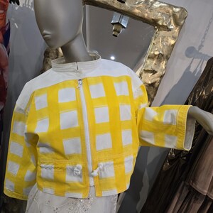 May include: A yellow and white checkered jacket with a white zipper and collar is displayed on a mannequin. The jacket has a cropped fit and drawstring details at the waist. The sleeves are three-quarter length.