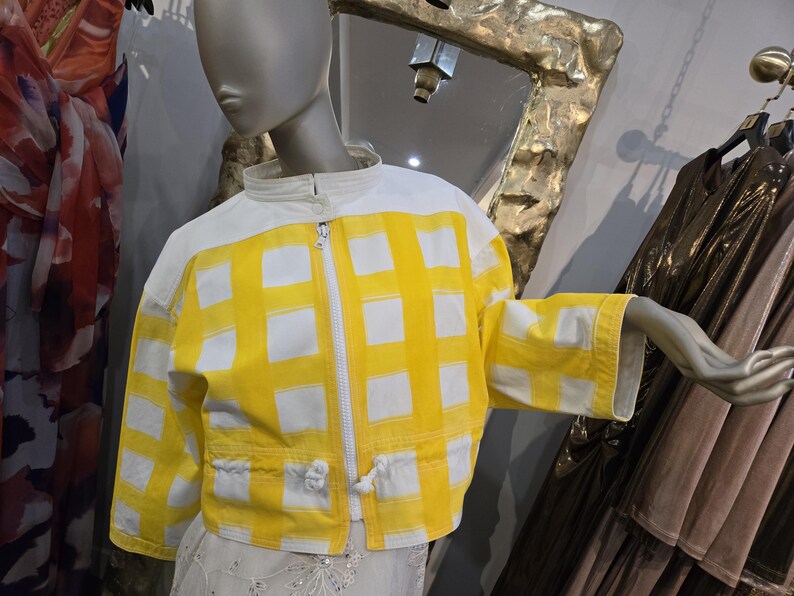 May include: A yellow and white checkered jacket with a white zipper and collar is displayed on a mannequin. The jacket has a cropped fit and drawstring details at the waist. The sleeves are three-quarter length.