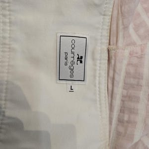 Może przedstawiać: Zbliżenie białego ubrania z metką Courr&egrave;ges Paris. Etykieta ma czarny tekst i logo. Rozmiar oznaczony jako "L". Tło obejmuje r&oacute;żowo-białą tkaninę w paski.