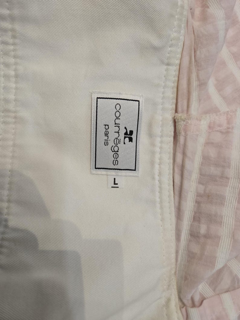 Może przedstawiać: Zbliżenie białego ubrania z metką Courr&egrave;ges Paris. Etykieta ma czarny tekst i logo. Rozmiar oznaczony jako "L". Tło obejmuje r&oacute;żowo-białą tkaninę w paski.