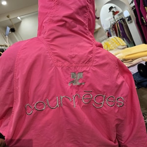 May include: Bright pink hooded jacket with the embroidered word "courr&egrave;ges" in silver thread across the back. The jacket has a silver logo above the brand name. The jacket is made of a lightweight material.