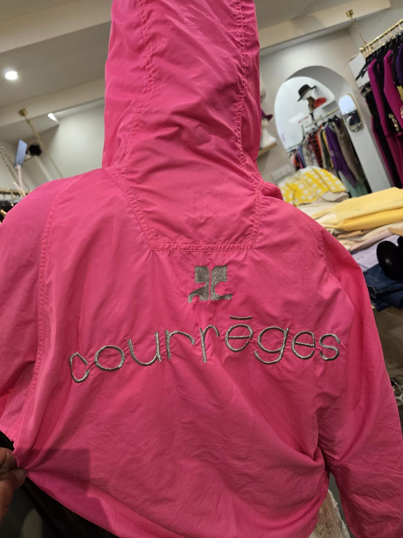 May include: Bright pink hooded jacket with the embroidered word "courr&egrave;ges" in silver thread across the back. The jacket has a silver logo above the brand name. The jacket is made of a lightweight material.
