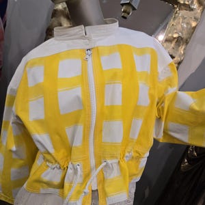 May include: A yellow and white jacket with a grid pattern. The jacket has a stand-up collar, a zipper closure, and a drawstring waist. The jacket is displayed on a mannequin.