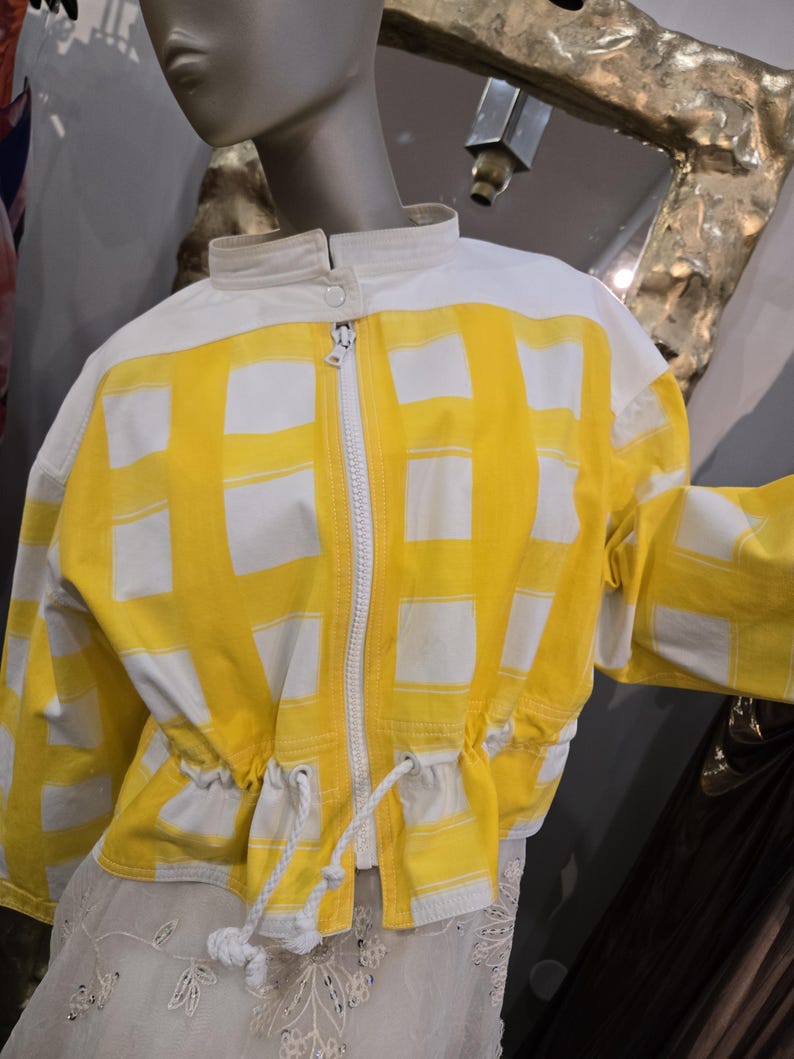 May include: A yellow and white jacket with a grid pattern. The jacket has a stand-up collar, a zipper closure, and a drawstring waist. The jacket is displayed on a mannequin.
