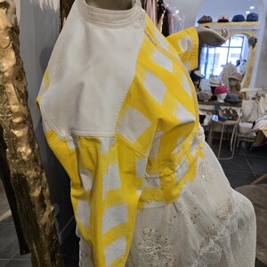 May include: A mannequin displays a white and yellow jacket with a geometric pattern. The jacket has a high collar and is paired with a white dress with floral embroidery and lace trim. The background shows a boutique setting.