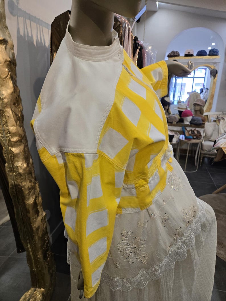 May include: A mannequin displays a white and yellow jacket with a geometric pattern. The jacket has a high collar and is paired with a white dress with floral embroidery and lace trim. The background shows a boutique setting.