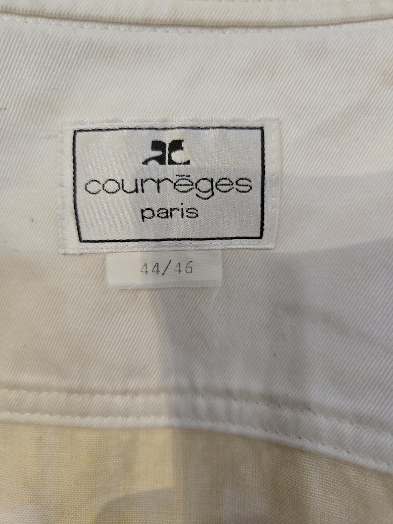 May include: Close-up of a white fabric tag with the text "courr&egrave;ges paris" in black, and the size "44/46" in black. The tag is rectangular with a black border and the Courr&egrave;ges logo. The fabric is a light cream color.