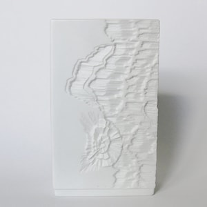 May include: White ceramic vase with a textured, raised design resembling a mountain range. The vase is tall and narrow, with a rectangular base.