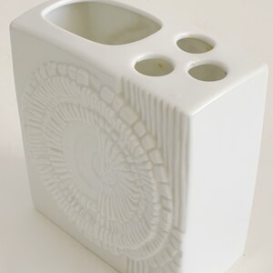Puede incluir: Un portacepillos de cer&aacute;mica blanca de forma rectangular. Cuenta con una gran abertura ovalada y tres peque&ntilde;os orificios circulares. El frente tiene un dise&ntilde;o circular texturizado en relieve. Un accesorio de ba&ntilde;o.