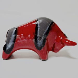 Peut inclure: Figurine de taureau en céramique rouge brillant avec des accents noirs et gris. La sculpture a un design stylisé, avec un corps incurvé et des cornes proéminentes. L'objet est décoratif.