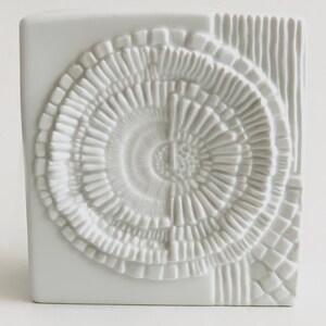 Puede incluir: Jarr&oacute;n de cer&aacute;mica blanca con forma cuadrada y un dise&ntilde;o circular texturizado en la parte delantera. El dise&ntilde;o presenta c&iacute;rculos conc&eacute;ntricos y l&iacute;neas verticales en el lateral. El jarr&oacute;n es un objeto decorativo.