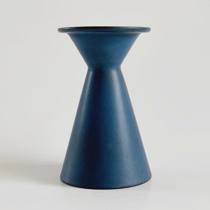 May include: A navy blue ceramic candle holder with a flared base and a narrow neck. The top is a flat, circular surface. The candle holder has a matte finish and a simple, modern design.