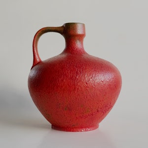 Puede incluir: Una jarra de cerámica roja con una superficie texturizada y una sola asa. La jarra tiene un cuerpo redondeado y un labio ligeramente acampanado.