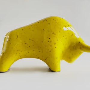 Könnte beinhalten: Eine Keramik-Stierfigur in leuchtendem Gelb, gesprenkelt mit dunkelbraunen Punkten. Die glänzende Oberfläche hebt die Form des Stiers mit seinen Hörnern und dem gewölbten Rücken hervor. Ein dekorativer Artikel für die Inneneinrichtung.