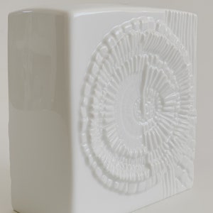 Puede incluir: Un jarr&oacute;n de cer&aacute;mica blanco con forma cuadrada. Presenta un dise&ntilde;o en espiral en relieve y l&iacute;neas verticales en el lado derecho. El jarr&oacute;n tiene un acabado brillante y est&aacute; destinado a la decoraci&oacute;n del hogar.
