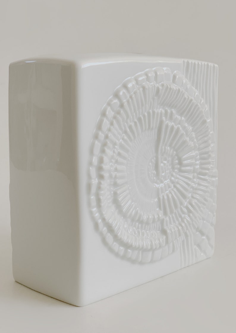 Puede incluir: Un jarr&oacute;n de cer&aacute;mica blanco con forma cuadrada. Presenta un dise&ntilde;o en espiral en relieve y l&iacute;neas verticales en el lado derecho. El jarr&oacute;n tiene un acabado brillante y est&aacute; destinado a la decoraci&oacute;n del hogar.