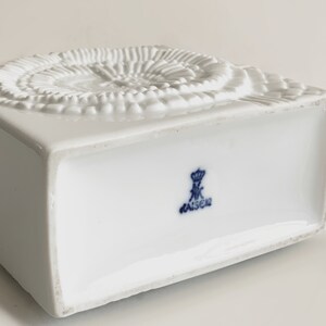 Puede incluir: Una caja de porcelana blanca con base cuadrada y dise&ntilde;o espiral texturizado en la parte superior. La parte inferior de la caja presenta un emblema azul con la palabra "KAISER".