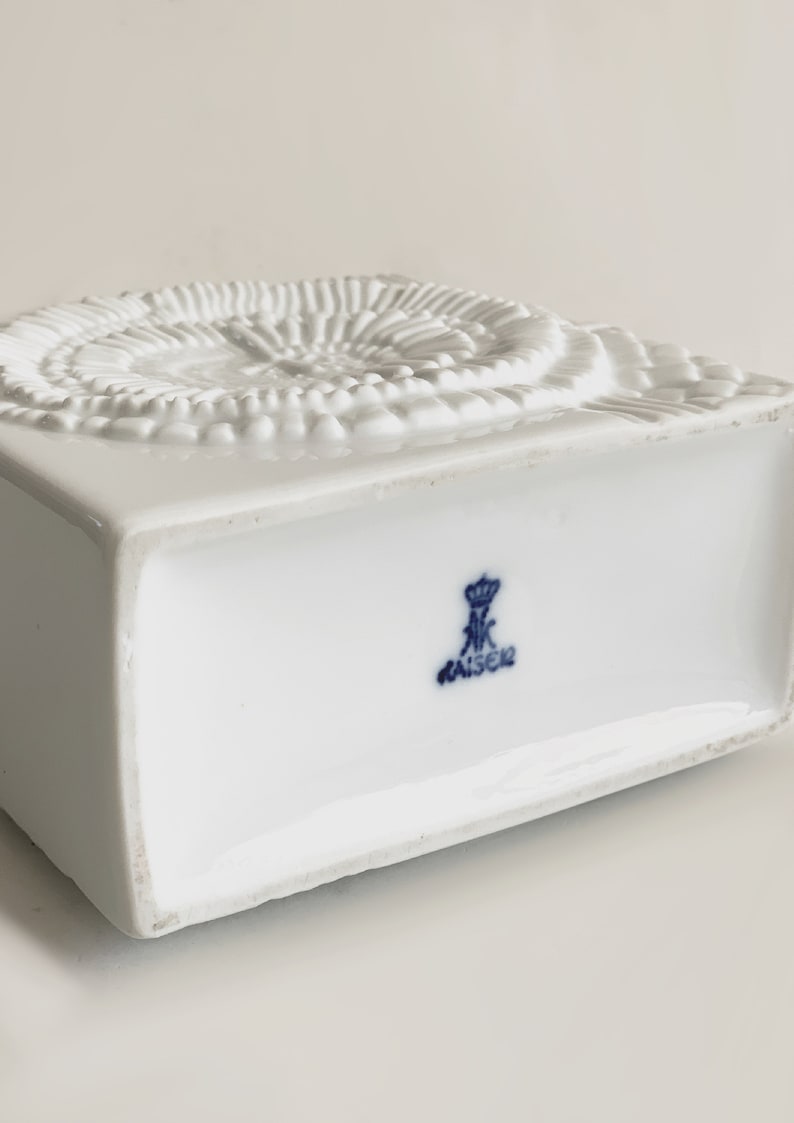 Puede incluir: Una caja de porcelana blanca con base cuadrada y dise&ntilde;o espiral texturizado en la parte superior. La parte inferior de la caja presenta un emblema azul con la palabra "KAISER".