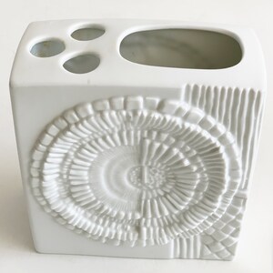 Puede incluir: Un portacepillos de cer&aacute;mica blanca con forma cuadrada y un dise&ntilde;o decorativo texturizado. El soporte tiene tres agujeros peque&ntilde;os y una abertura m&aacute;s grande en la parte superior. La parte delantera presenta un patr&oacute;n circular en espiral y el lateral l&iacute;neas verticales.
