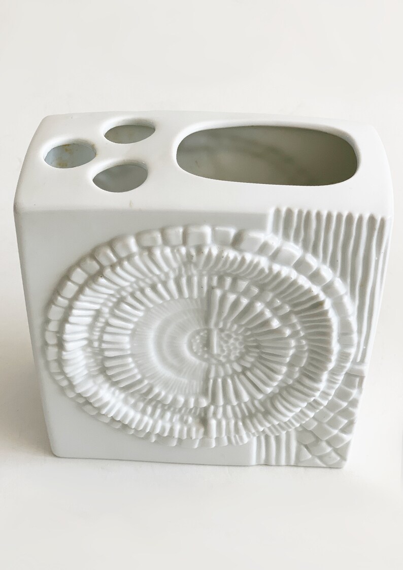 Puede incluir: Un portacepillos de cer&aacute;mica blanca con forma cuadrada y un dise&ntilde;o decorativo texturizado. El soporte tiene tres agujeros peque&ntilde;os y una abertura m&aacute;s grande en la parte superior. La parte delantera presenta un patr&oacute;n circular en espiral y el lateral l&iacute;neas verticales.